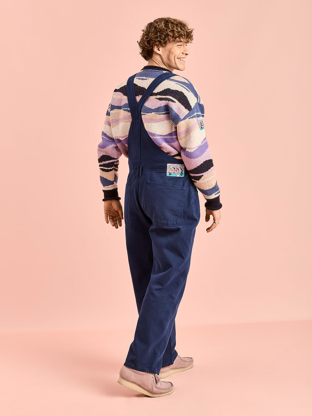 Male Model wears the navy overall style dungarees, showing the two back pockets and the branded seasonal badge on the waistband 